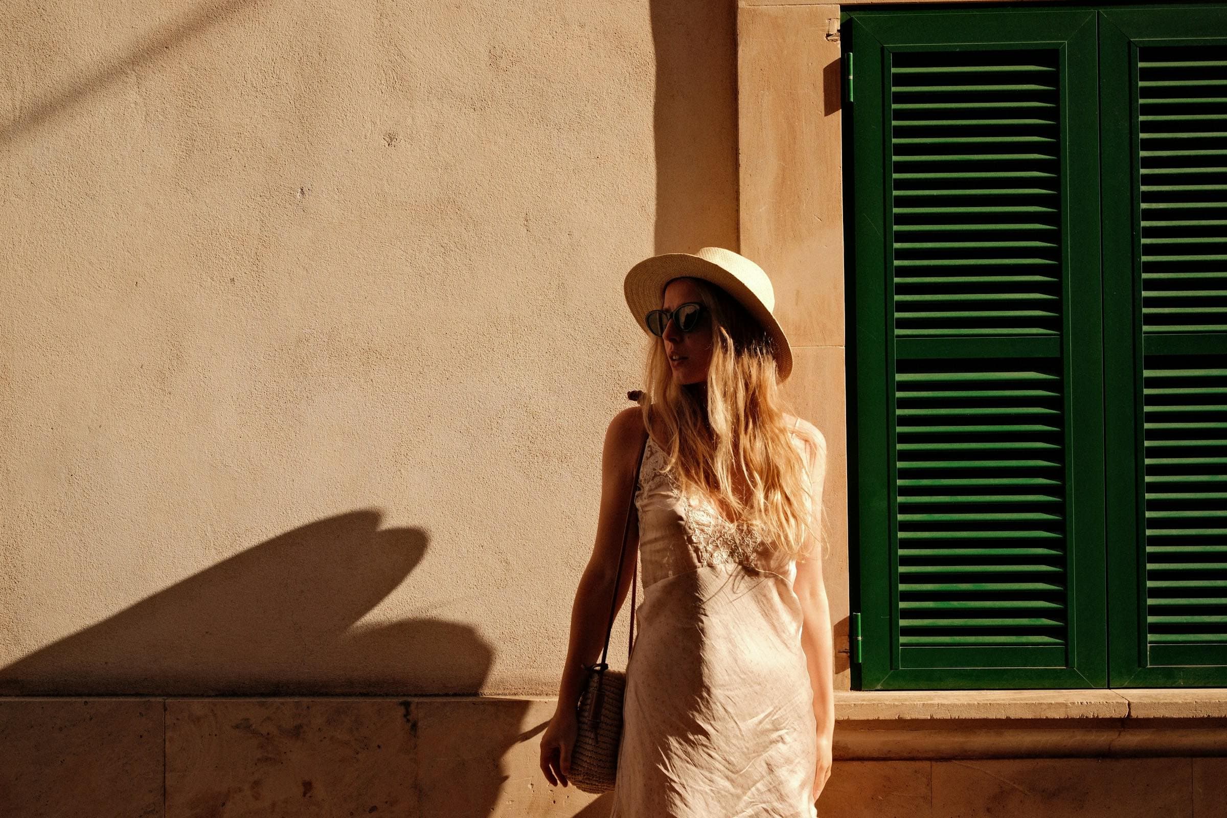 A woman in a straw hat and dark sunglasses against a sunlit wall with green shutters